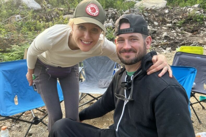 a young couple jill and sean leary are in a campsite setting sean is seated in his chair and jill crouches next to him with her arm around his shoulders