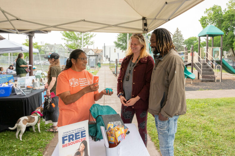 at an outdoor community fair, a native american woman in an FFN tee shirt hands sensory toys to a pregnant woman and her partner. a sign reading free resources sits on the table.